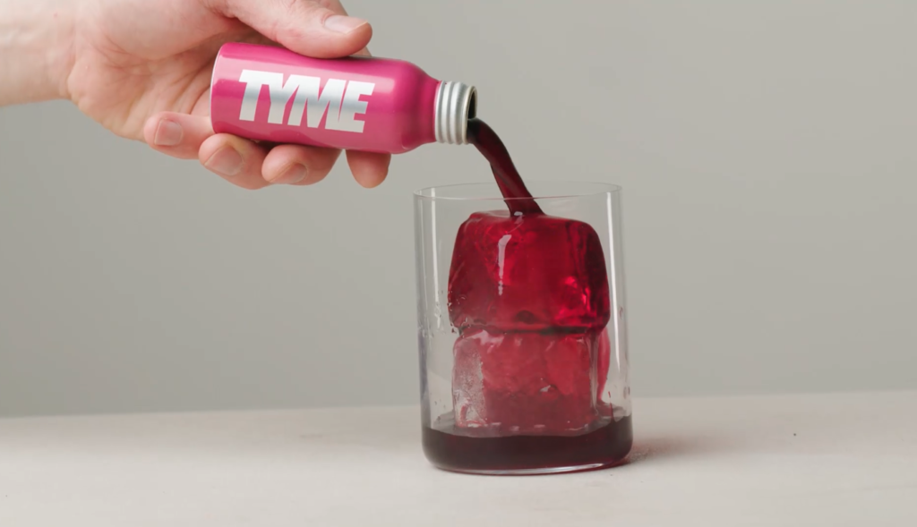 Small pink bottle of Tyme's Elderberry and Beetroot shot being poured over ice into a glass tumbler.